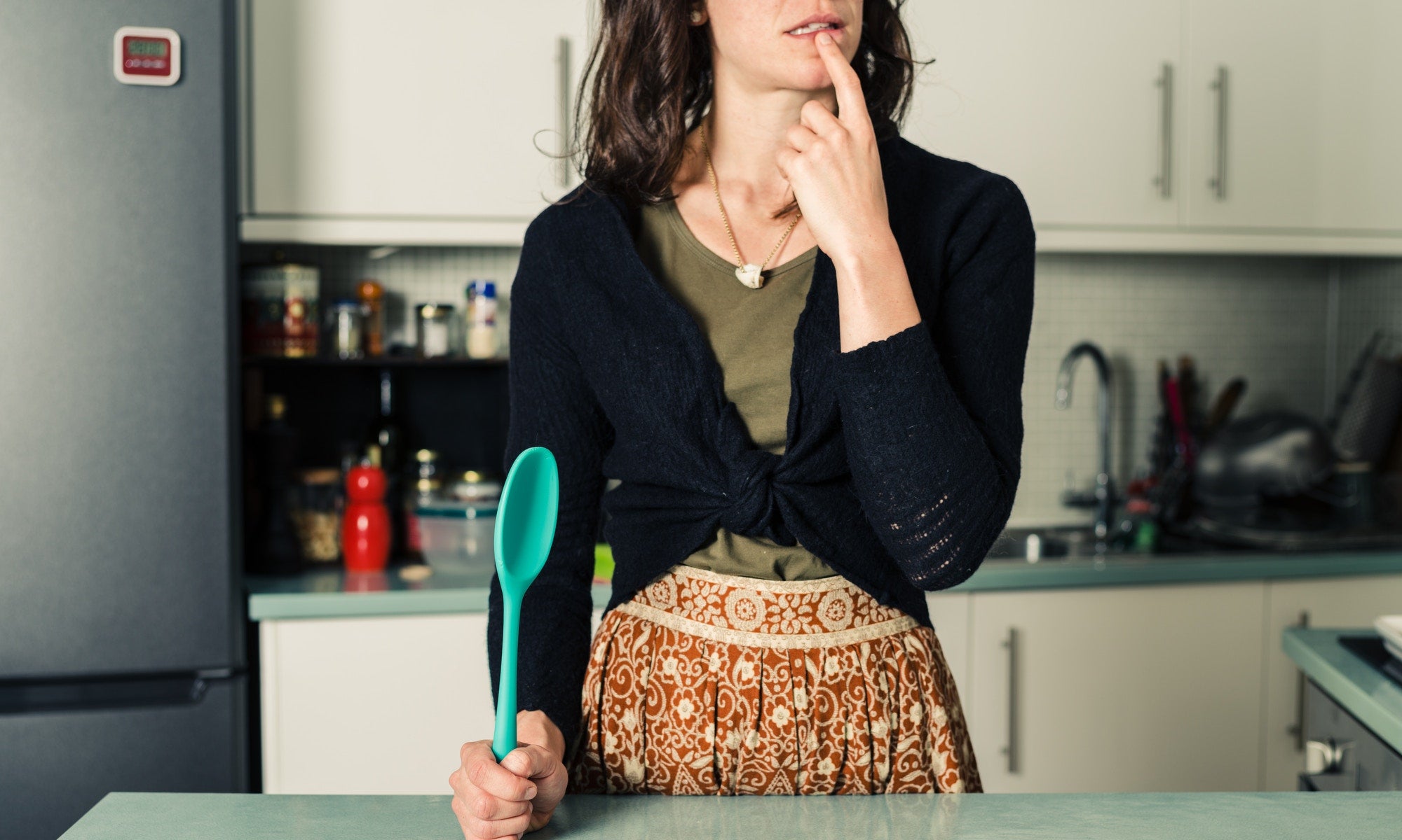 Confused woman with spoon in kitchen.