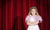 Front view of young girl standing in spotlight against red stage curtain looking annoyed