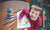 little boy looking at camera, smiling while painting picture sitting at table in art class