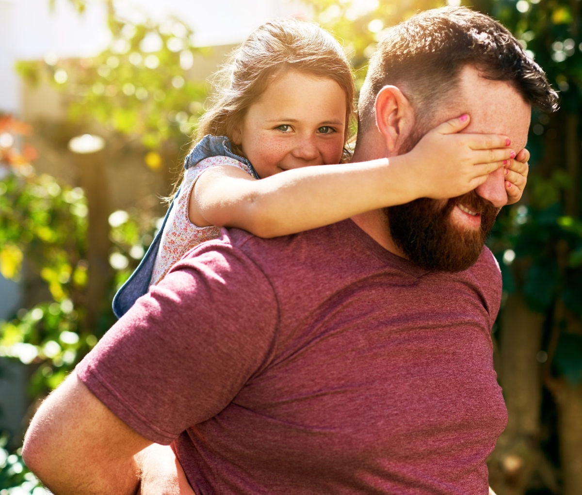 little girl piggy riding on her father closing his eyes with her hand.