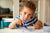 young boy drawing with crayons on sheets of white paper on a table