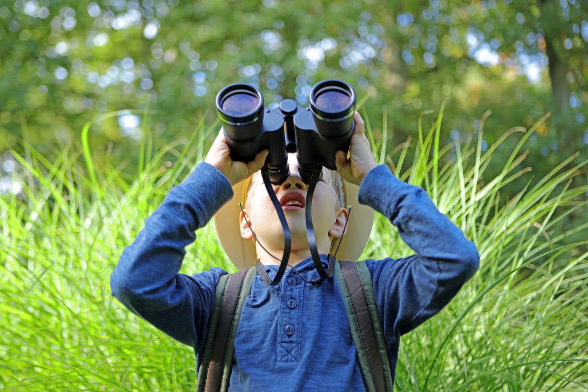 Boy in pith helmet looking up through big binoculars outdoors